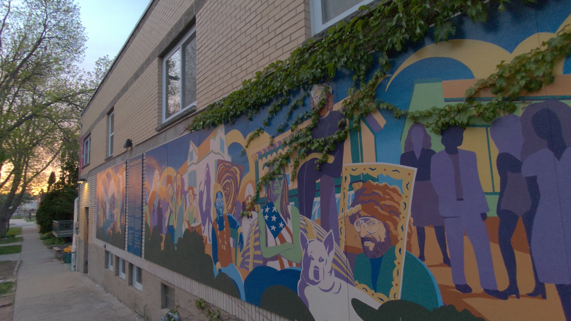 A brick building at dusk with a long, brightly colored community mural of painted figures, a dog and an American flag along its wall under trailing ivy, shown in full color as a sample of the camera's passthrough view.